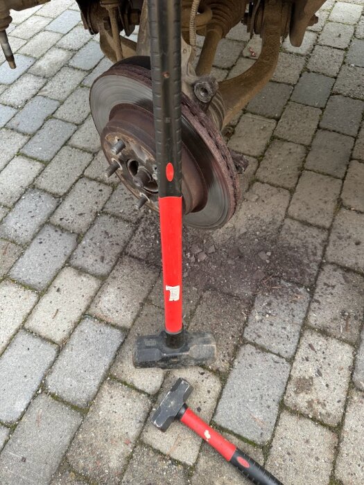 Två hammare, en större och en mindre, ligger på marken bredvid ett bilhjul utan däck på stenläggning.