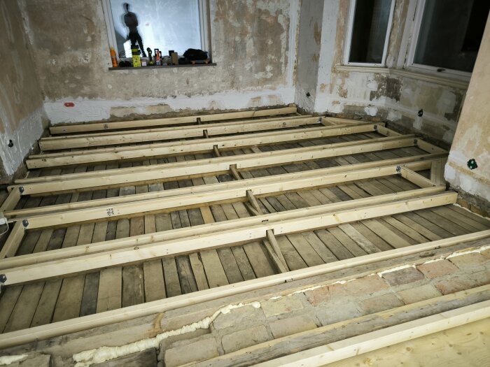 Rum under renovering med synliga golvbjälkar och fogskum i springorna, samt verktyg och material vid ett fönster.