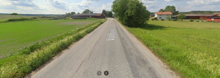 Landsbygdsväg i tätort omgiven av gröna fält och lantliga byggnader under en blå himmel med lätta moln.