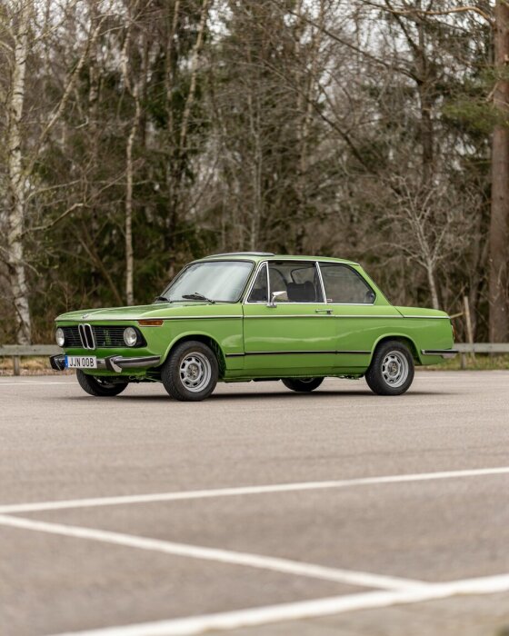 Grön vintagebil parkerad på tom parkering, med träd i bakgrunden.