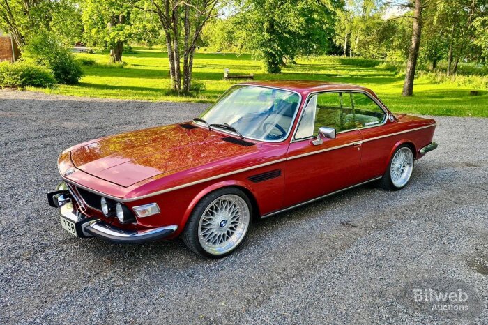 Röd klassisk bil modell BMW från München, parkerad på grusväg med grönskande natur i bakgrunden.