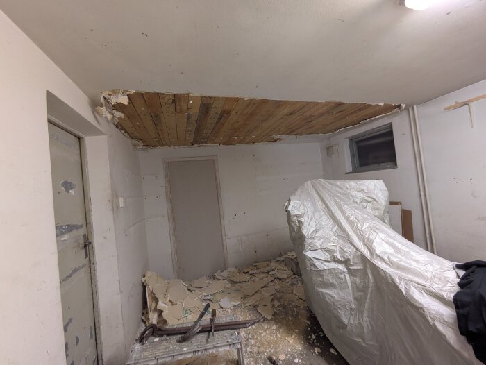 Room under renovation with partially removed ceiling, tools on the floor, debris scattered, and a large covered object in the corner.