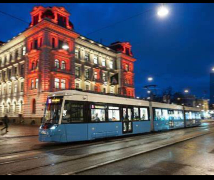 Blå spårvagn kör förbi ett upplyst, historiskt byggnad med röd belysning i kvällsmörker.