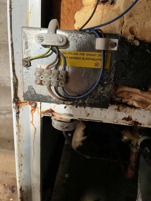 Elanslutning med kablar och terminalblock vid en varmvattenberedare. Skylt säger "inkoppling får endast göras av behörig elinstallatör".