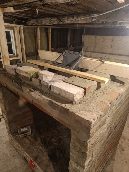 En ofärdig murad öppen spis under konstruktion med ett vridspjäll och trästycken ovanpå strukturen i ett byggprojekt.