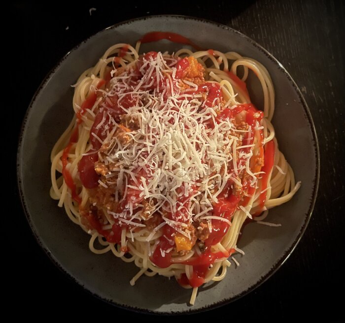 Tallrik med spaghetti och köttfärssås toppad med riven ost och ketchup, serverad på mörk yta.