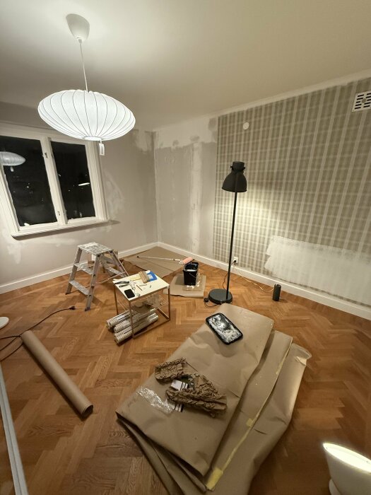 Sovrum under renovering med trägolv, stege, arbetsbord med verktyg, golvlampa och förpackningsmaterial. Vägg renoveras och delvis tapetseras.