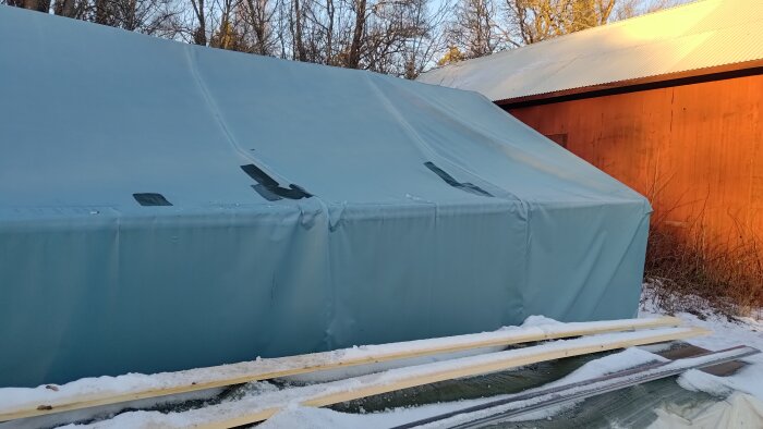 Tält med reparerad presenning i snöigt landskap intill en röd byggnad, med plankor i förgrunden.