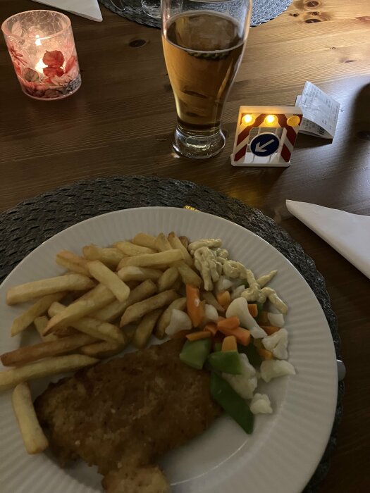 Tallrik med schnitzel, pommes frites, frysta grönsaker och remouladsås, glas öl, tänd ljuslykta och en skojig mini-vägskylt på ett bord.