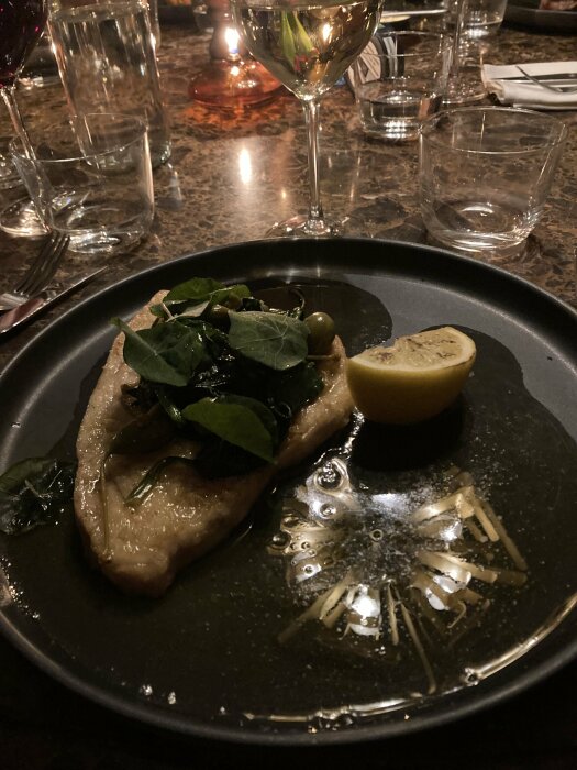 En tallrik med eldad plattfisk dekorerad med gröna blad och citronklyfta, serverad med ett glas vitt vin på ett marmorbord.