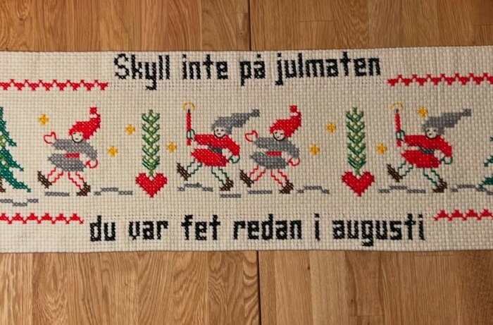 Broderad löpare med texten "Skyll inte på julmaten, du var fet redan i augusti" och motiv av skridskoåkande figurer.