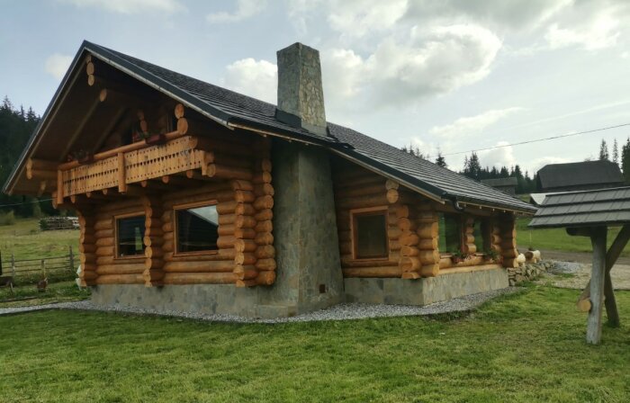 A log house with a stone chimney and steel roof, set in a grassy landscape, illustrating a prefabricated home for potential import and assembly.