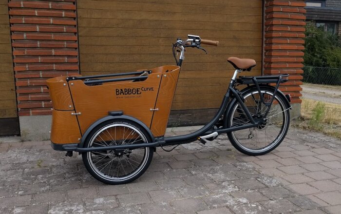 Lastcykel av märket Babboe Curve, parkerad framför en tegelvägg.