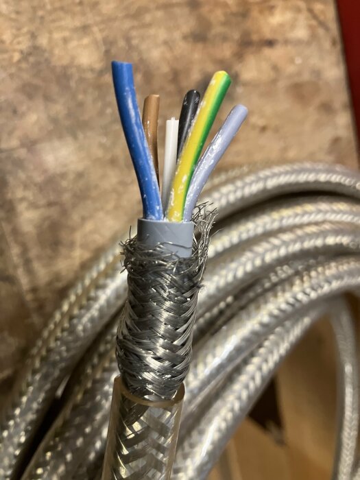 Flexibel anslutningskabel med metallstrumpa och synliga färgade ledare på träbord, används för rörliga maskiner.