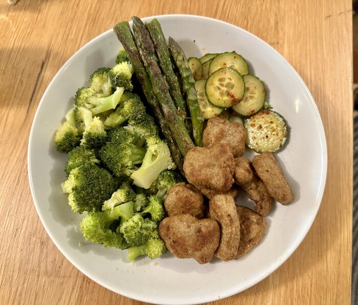 Tallrik med kycklingbitar, broccoli, sparris och skivad zucchini pÄ trÀbord.