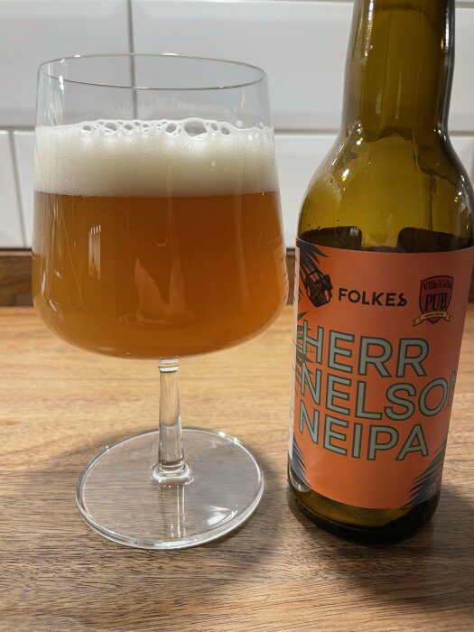 En glas med öl bredvid en flaska märkt "Herr Nelson NEIPA" av märket Folkes, står på ett träbord.