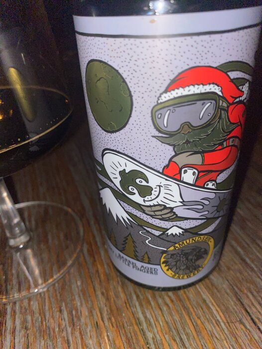 Flaska av Amundsen Sticka little fingers Barrel aged Stout med vintertema, bredvid ett vinglas på ett trägolv.