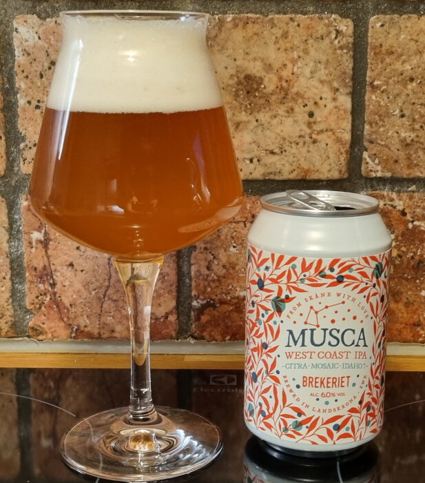 Ölglas fyllt med bärnstensfärgad West Coast IPA bredvid Musca ölburk, mot bakgrund av tegelvägg.