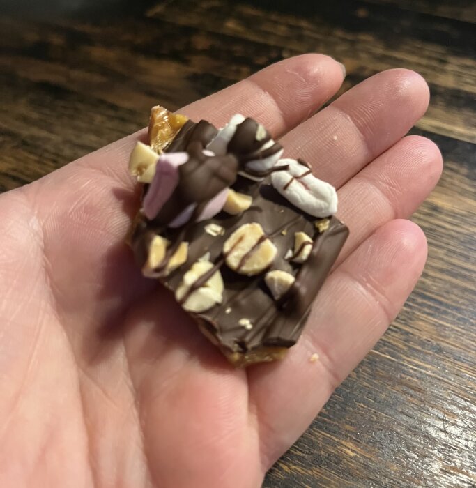 Hand håller en bit av hemmagjord rocky road med marshmallows, choklad och nötter på ett träbord.