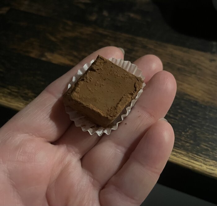Hand håller en bit chokladtryffel i ett rynkigt papper på ett mörkt bord.