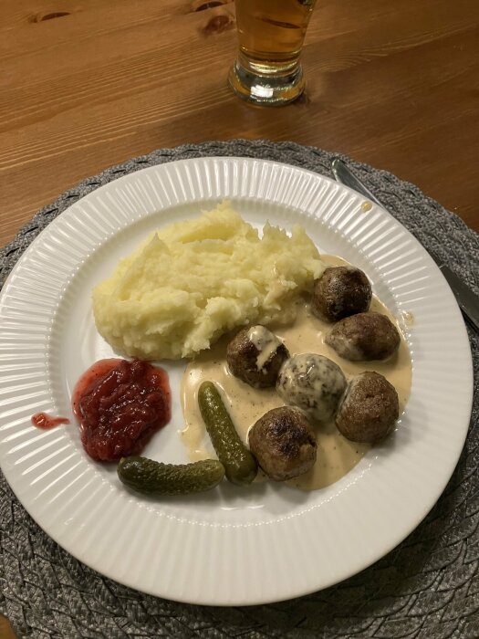 Tallrik med köttbullar i gräddsås, potatismos, lingonsylt och cornichons på ett bord.