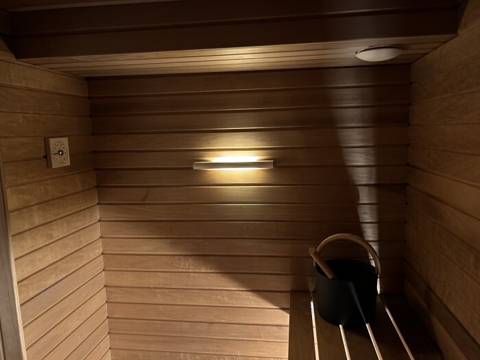 Inre bastuvägg med trälavar och en träbänk, en vägglampa som lyser och en svart hink med trähandtag.