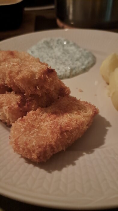 Panko-panerade torskbitar med potatisskivor och en krämig, vit sås på en tallrik.