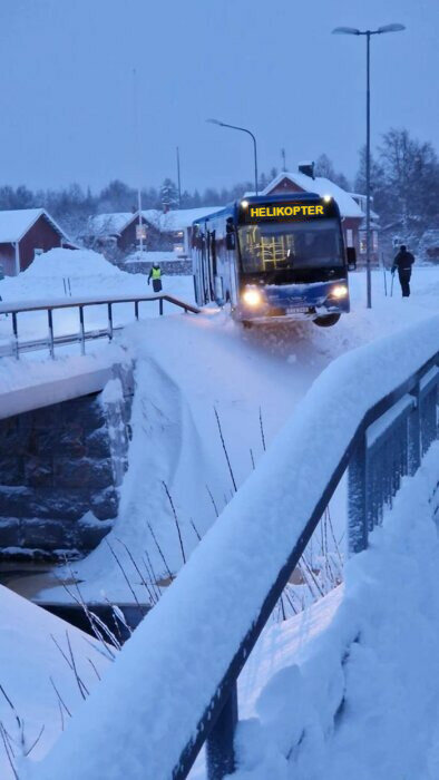 Buss i vinterlandskap med texten "HELIKOPTER" på displayen kör på snötäckt väg bredvid snötyngda hus och gångväg.