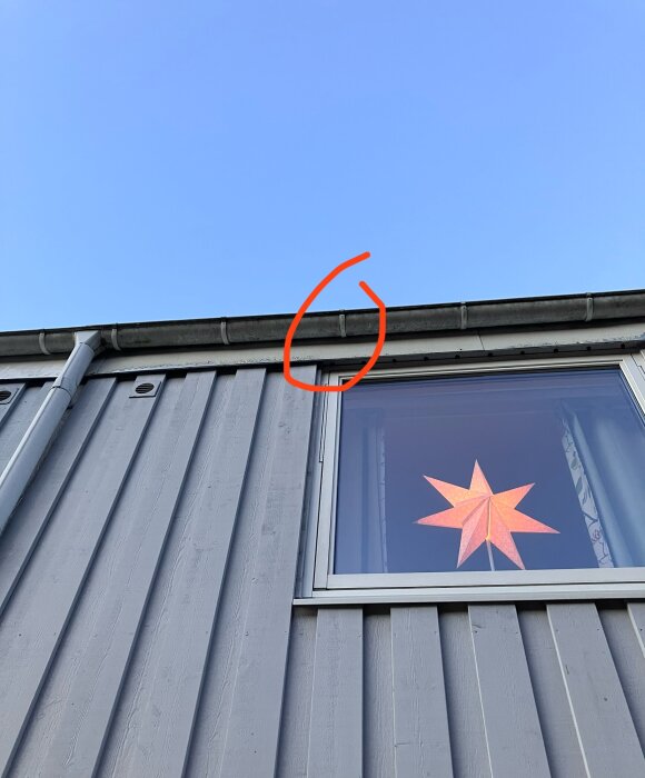 Takränna på hus med fönster och stjärnformad lampa inuti, röd cirkel markerar område på taket under blå himmel.