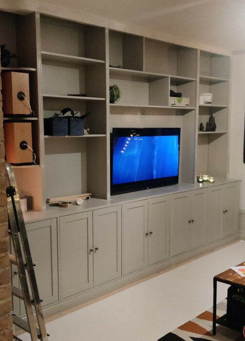 Grå hylla under byggnation med TV i mitten och förvaringshyllor runtom. Verktyg, stege och dekorativa föremål syns på hyllorna.