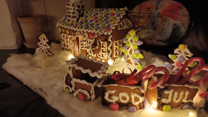 Pepparkakshus dekorerat med glansigt godis, ljusslingor och texten "God Jul" i en julig miljö.