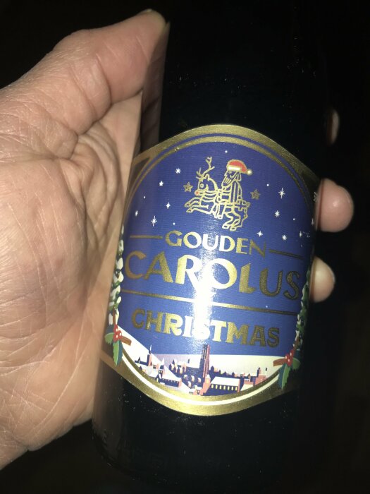 En hand håller en flaska Gouden Carolus Christmas öl med etikett i juldesign.