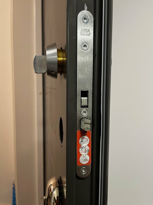 Närbild på en dörr med ett ASSA-lås i demontering. Låskolven och skruvar synliga, med instruktioner på en orange etikett.