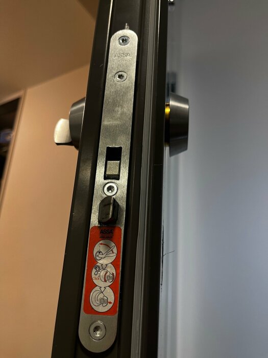 Närbild på dörrkantslås med instruktioner för demontering, synlig spärr och dekal från ASSA.