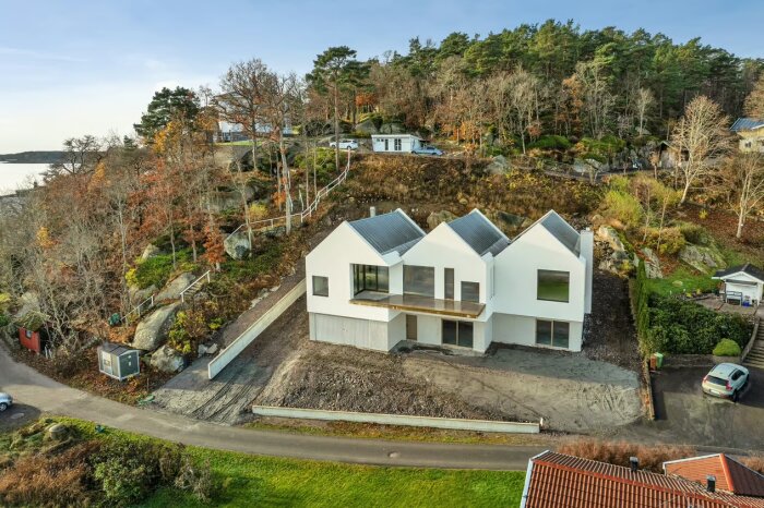 Modern villa med vit fasad och stora fönster, omgiven av träd och skog på en sluttning vid havet, med en uppfart i Kungsbacka.