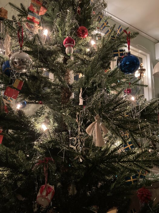 Julgran dekorerad med ljus, glittrande kulor, små svenska flaggor och hemmagjorda prydnader. Granen ger en traditionell och festlig känsla.