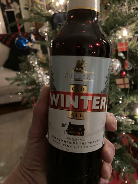 Person håller en flaska Fuller’s Old Winter Ale framför en pyntad julgran.