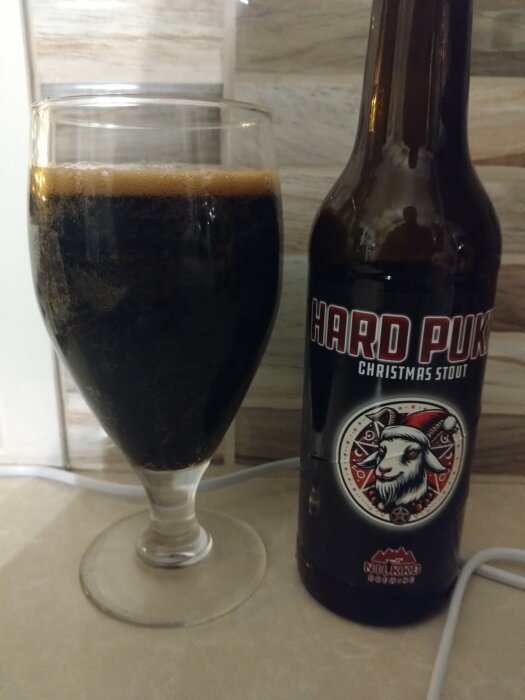 Flaska och glas av mörk, skummande öl med etiketten "Hard Pukki Christmas Stout" från NUKKOpanimo, fotograferad mot kakelvägg.