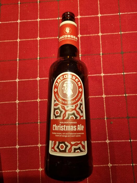 Flaska med Jacobsen Christmas Ale, 75 cl, placerad på röd bordsduk. Etykett i rött och guld med beskrivning av smakprofil.