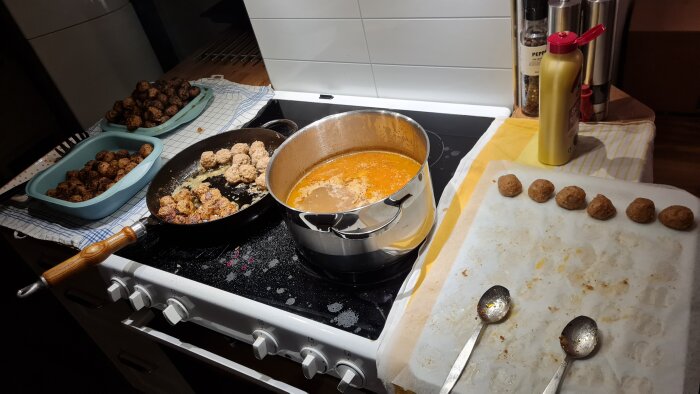 Kök med hemgjorda köttbullar i stekpanna, kastrull och på bakplåtspapper. Två skålar med klara köttbullar och en flaska senap bredvid.