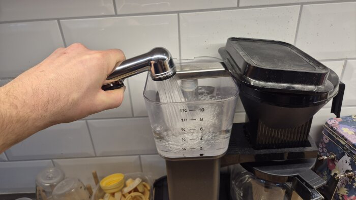 Hand håller i en duschslang och fyller en behållare med vatten bredvid en kaffebryggare i köket.