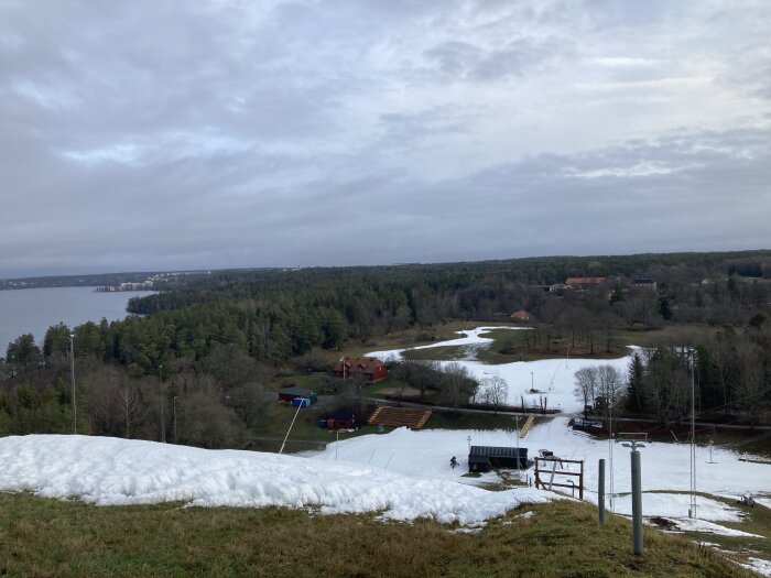 Vintrig landskapsvy med snötäckt skidbacke, omgiven av skog och gräs, med molnig himmel över en sjö och avlägsna byggnader i bakgrunden.