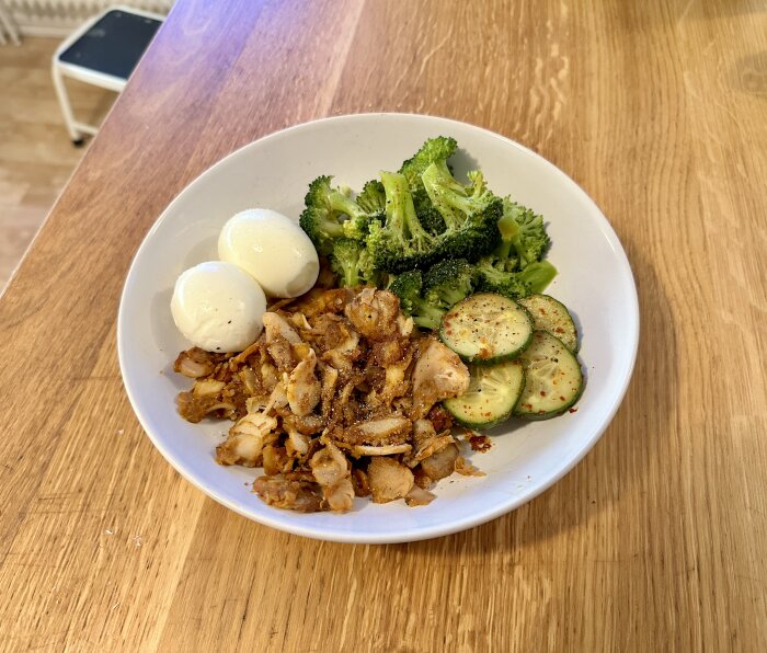 Skål med kokta ägg, broccoli, skivad gurka och kryddat kycklingkött på ett träbord.