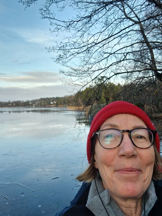 Person i röd mössa och glasögon framför en frusen sjö med träd i bakgrunden under en klar himmel.