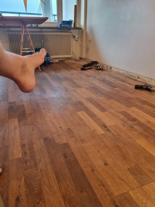 Trägolv under renovering med verktyg och borttagna lister. Synlig radiator och bord vid fönstret i bakgrunden. En fot skymtar i hörnet.