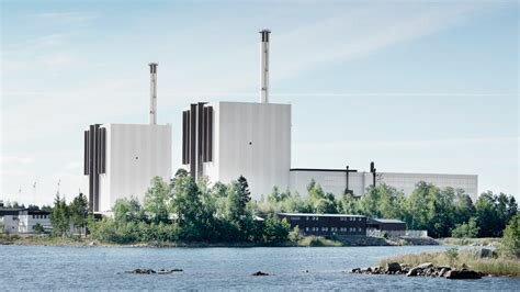 Två stora vita industribyggnader med höga skorstenar vid en sjö, omgivna av grönska och blå himmel.