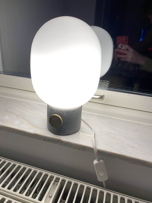 Bordslampa med vit skärm på fönsterbräda, ansluten till en strömbrytare istället för en dimmer, med en mässingsknapp på lampbasen.