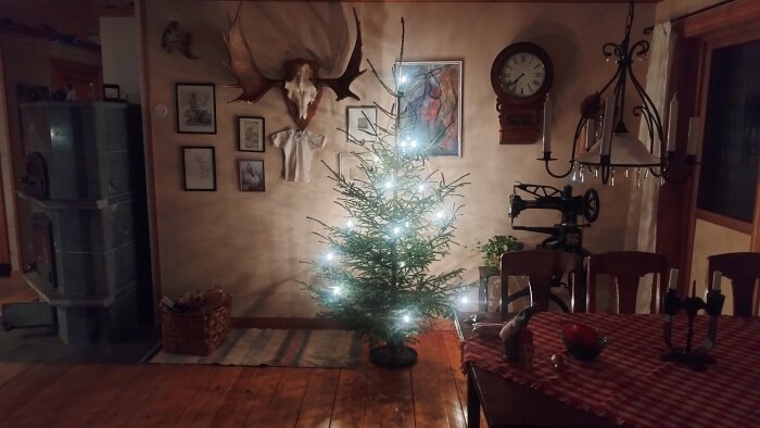 Julgran med ljus i ett rustikt rum med trägolv, eldstad, matbord med röd duk, gammal symaskin och väggdekorationer, inklusive ett älghorn och klocka.