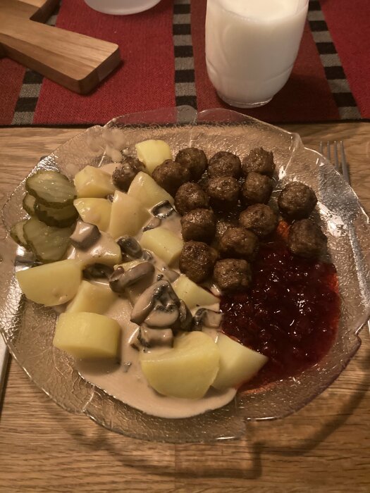 Tallrik med delikatessköttbullar, champinjonsås, potatis, inlagd gurka och lingonsylt. Glas med mjölk i bakgrunden.