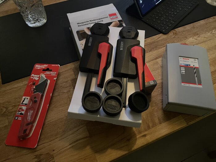 Paket med två Bessey-spännare, Milwaukee fällkniv, och Bosch borrset på ett träbord bredvid en surfplatta.